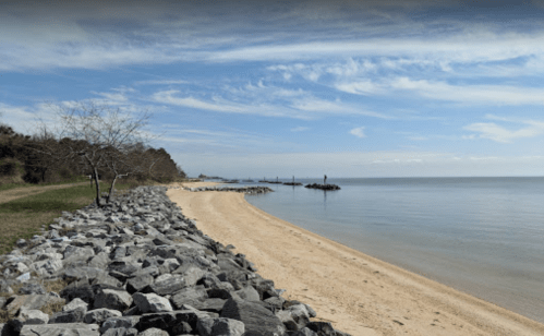 A serene beach scene with a sandy shore, rocky coastline, and calm water under a blue sky with scattered clouds.