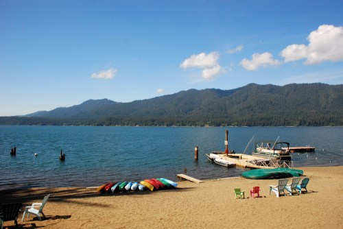 A serene lakeside view with colorful kayaks on the sand, a dock, and mountains in the background under a clear blue sky.