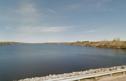 A calm river scene with gentle hills and a clear blue sky, bordered by a rocky shoreline.