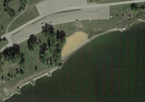 Aerial view of a sandy beach area next to a lake, with a parking lot and trees nearby.