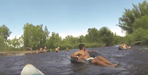 A group of people floating on inner tubes in a river, surrounded by trees and a clear blue sky.