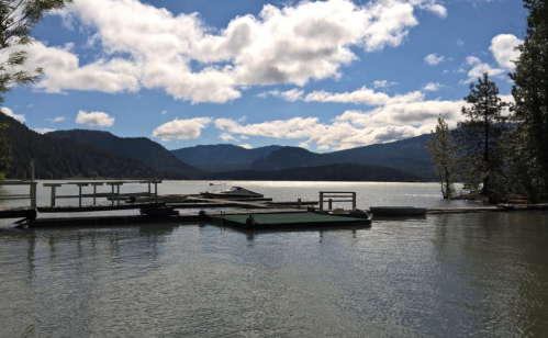A serene lake scene with a wooden dock, surrounded by mountains and trees under a partly cloudy sky.