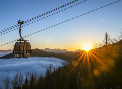A cable car glides above a sea of clouds at sunrise, with mountains silhouetted in the background.
