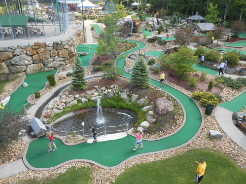 Aerial view of a mini-golf course with winding paths, a pond, and players enjoying the game amidst greenery.