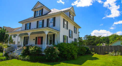 A charming white two-story house with a porch, surrounded by greenery and blue skies.