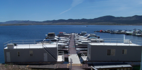 A marina with boats docked along a wooden pier, surrounded by calm water and mountains in the background.