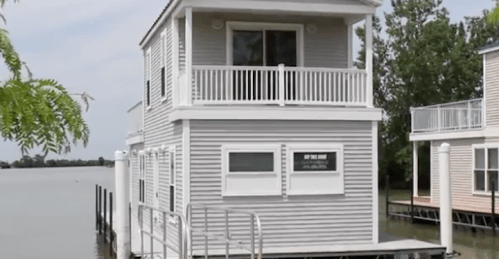 A two-story houseboat with a balcony, situated on calm water, surrounded by trees and other similar structures.