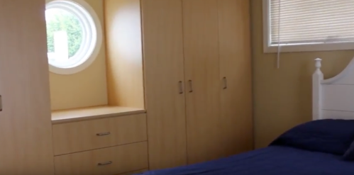 A cozy bedroom featuring a round window, wooden cabinets, and a neatly made bed with blue bedding.