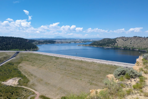 A scenic view of a lake surrounded by hills and trees under a blue sky with scattered clouds.