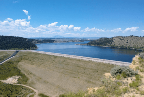 A scenic view of a lake surrounded by hills and trees under a blue sky with scattered clouds.