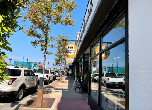 A sunny street scene featuring shops, trees, and parked cars along a sidewalk with a sign for a business.