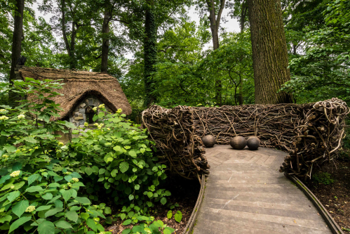 A rustic wooden path leads to a unique, woven structure surrounded by lush greenery and a small stone cottage.