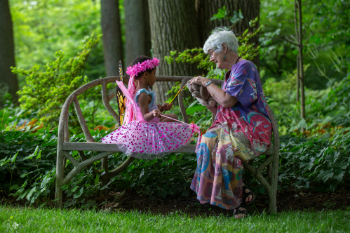A young girl in a pink dress with wings sits on a bench, chatting with an elderly woman in a colorful outfit.