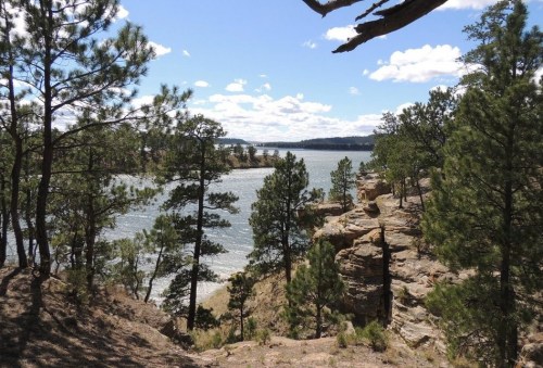 A scenic view of a lake surrounded by rocky cliffs and pine trees under a partly cloudy sky.