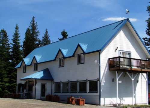 A large white house with a blue metal roof, surrounded by trees and featuring a balcony and flower boxes.