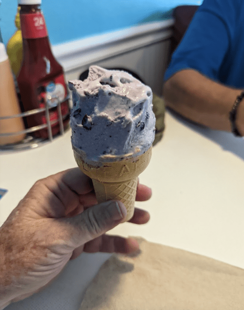A hand holding a cone of blueberry ice cream with chocolate chips, set on a table with a blurred background.