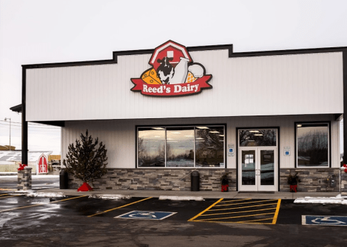 Exterior of Reed's Dairy store, featuring a sign with a cow logo, large windows, and a parking lot.