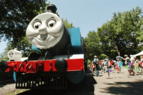 A smiling blue train with a friendly face, surrounded by a crowd of people in a park setting.