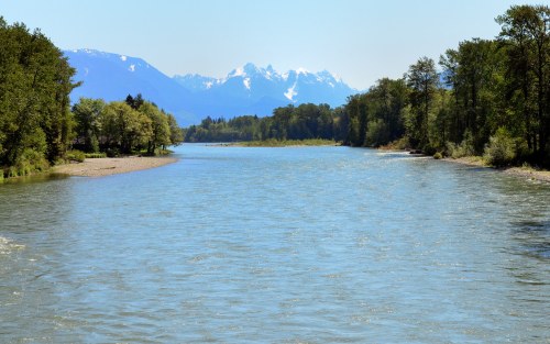 A serene river flows through a lush landscape, with snow-capped mountains in the background under a clear blue sky.