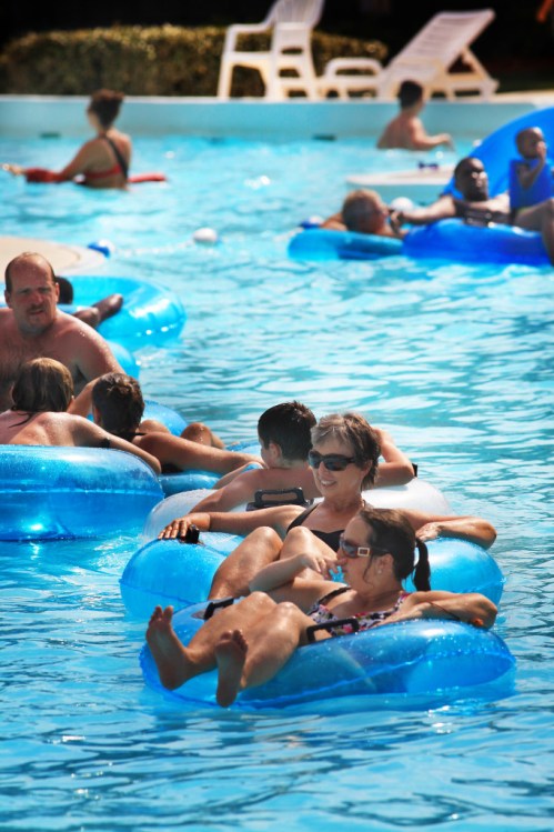 People relaxing on blue inner tubes in a pool, enjoying a sunny day with others in the background.