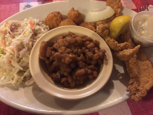 A plate of fried fish, coleslaw, baked beans, hushpuppies, onion slices, and a lemon wedge.
