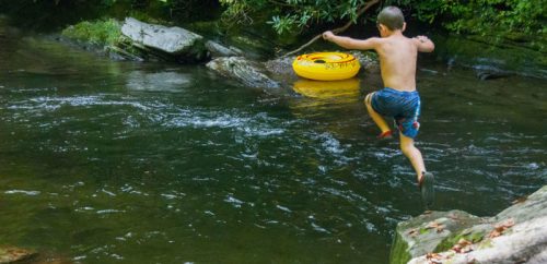 A boy jumps into a creek near a yellow inner tube, surrounded by lush greenery and rocky banks.