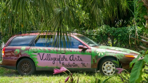 A colorful, painted car with "Village of the Arts, Bradenton, FL" written on it, surrounded by greenery.