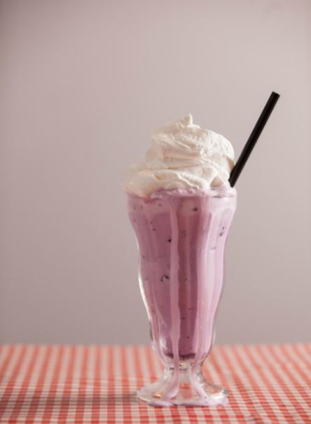 A tall glass filled with purple milkshake topped with whipped cream and a black straw, set on a checkered tablecloth.