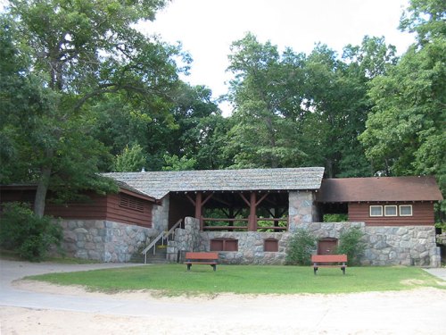 A rustic stone and wood pavilion surrounded by trees, with benches and a grassy area in front.