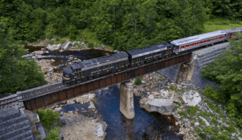 A train crosses a bridge over a river, surrounded by lush greenery and rocky terrain.