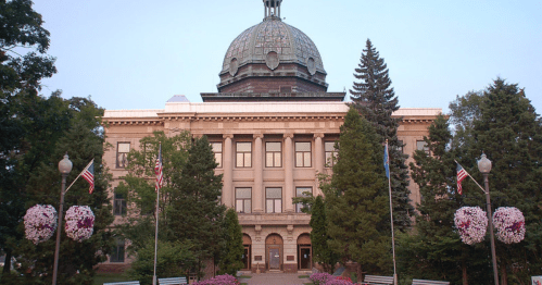 Historic building with a dome, surrounded by trees and flowers, featuring flags and a welcoming entrance.