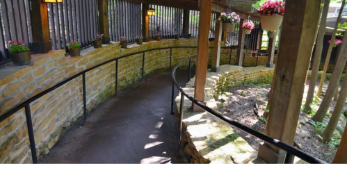 A winding pathway with a stone wall, surrounded by wooden beams and hanging flower pots in a serene setting.