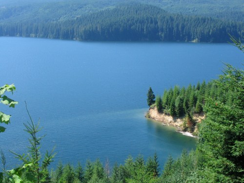 A serene lake surrounded by lush green forests and a small sandy peninsula extending into the water.