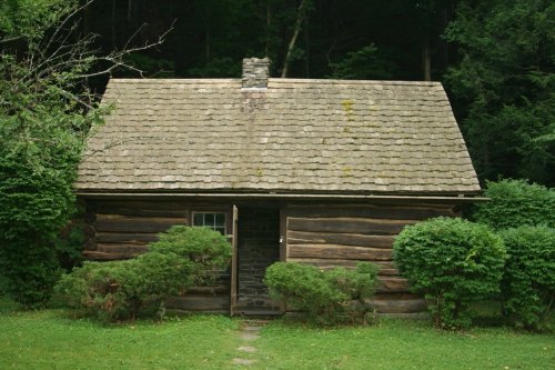 A rustic log cabin with a stone chimney, surrounded by greenery and trees, set in a peaceful natural environment.