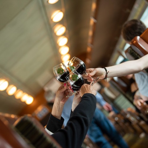 Two hands clink glasses of red wine in a cozy, well-lit setting, with people in the background enjoying their time.