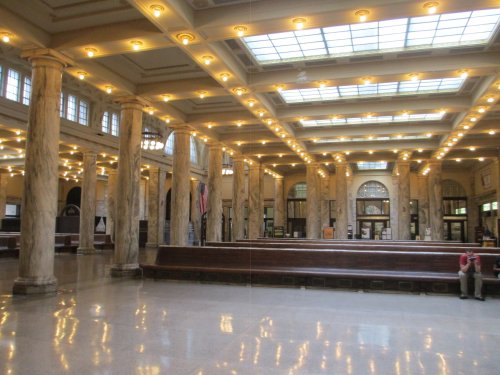 Interior of a grand hall with marble columns, bright lights, and a spacious layout, featuring wooden benches and flags.