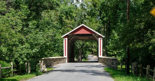 A red and white covered bridge spans a tree-lined road, surrounded by lush greenery.