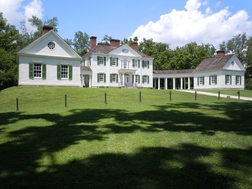 A large, white historic house with green lawns and trees under a blue sky, featuring a porch and multiple chimneys.