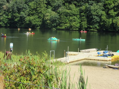 A serene lake scene with people kayaking and paddle boating, surrounded by lush greenery and a sandy shore.