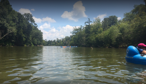 A serene river scene with lush greenery on both sides and a person in a blue float, enjoying a sunny day.