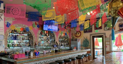 Colorful papel picado decorations hang above a vibrant bar with shelves of liquor and a lively atmosphere.