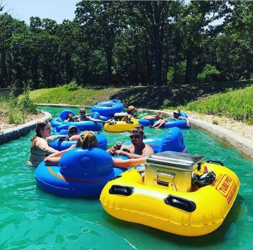 A group of people relaxes in blue and yellow inner tubes on a lazy river surrounded by greenery.