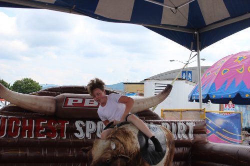 A child rides a mechanical bull at a fair, with a tent overhead and colorful decorations in the background.
