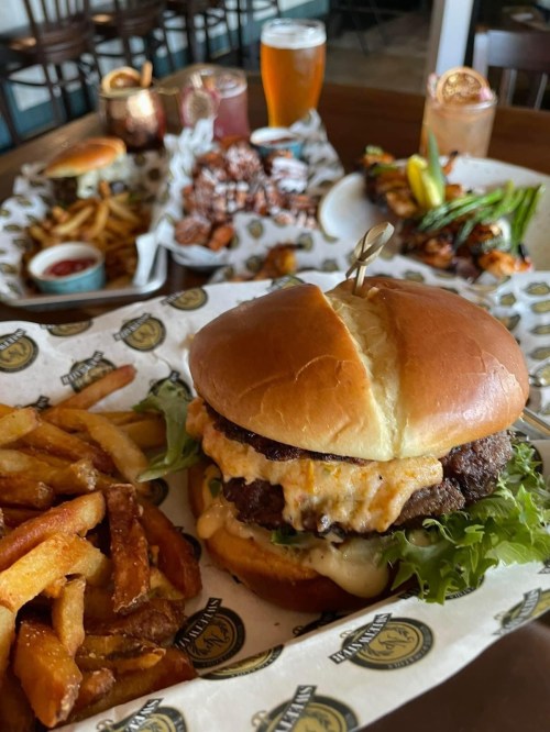 A close-up of a burger with cheese and lettuce, served with fries, alongside a variety of appetizers and drinks in the background.