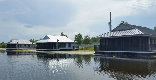 Three modern houses on stilts by a calm water body, surrounded by greenery under a cloudy sky.