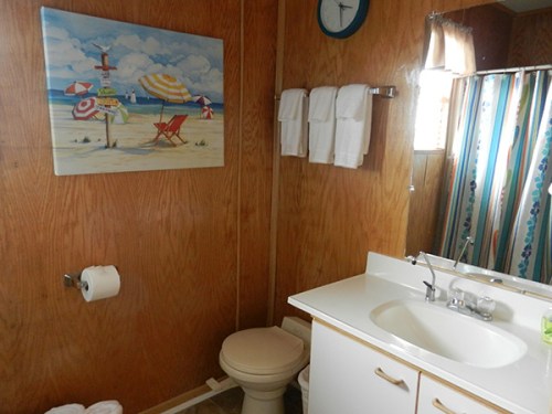 A bathroom with wooden walls, a beach-themed painting, towels, a toilet, and a sink with a mirror.