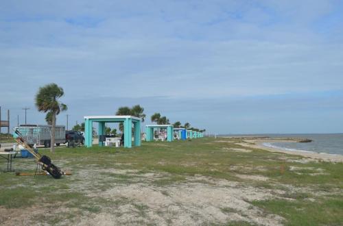 Coastal scene with turquoise structures along a grassy shore, palm trees, and a calm sea under a cloudy sky.