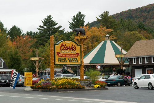 Sign for Clark's Trading Post with a bear statue, surrounded by colorful autumn trees and parked cars.
