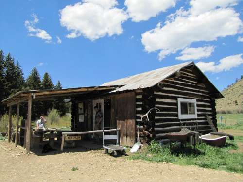 A rustic log cabin with a porch, surrounded by trees and blue skies, featuring a person sitting at a table outside.