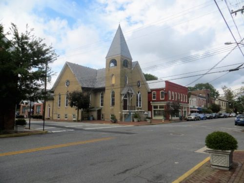 A quaint street scene featuring a church with a tall steeple and nearby red brick buildings under a cloudy sky.